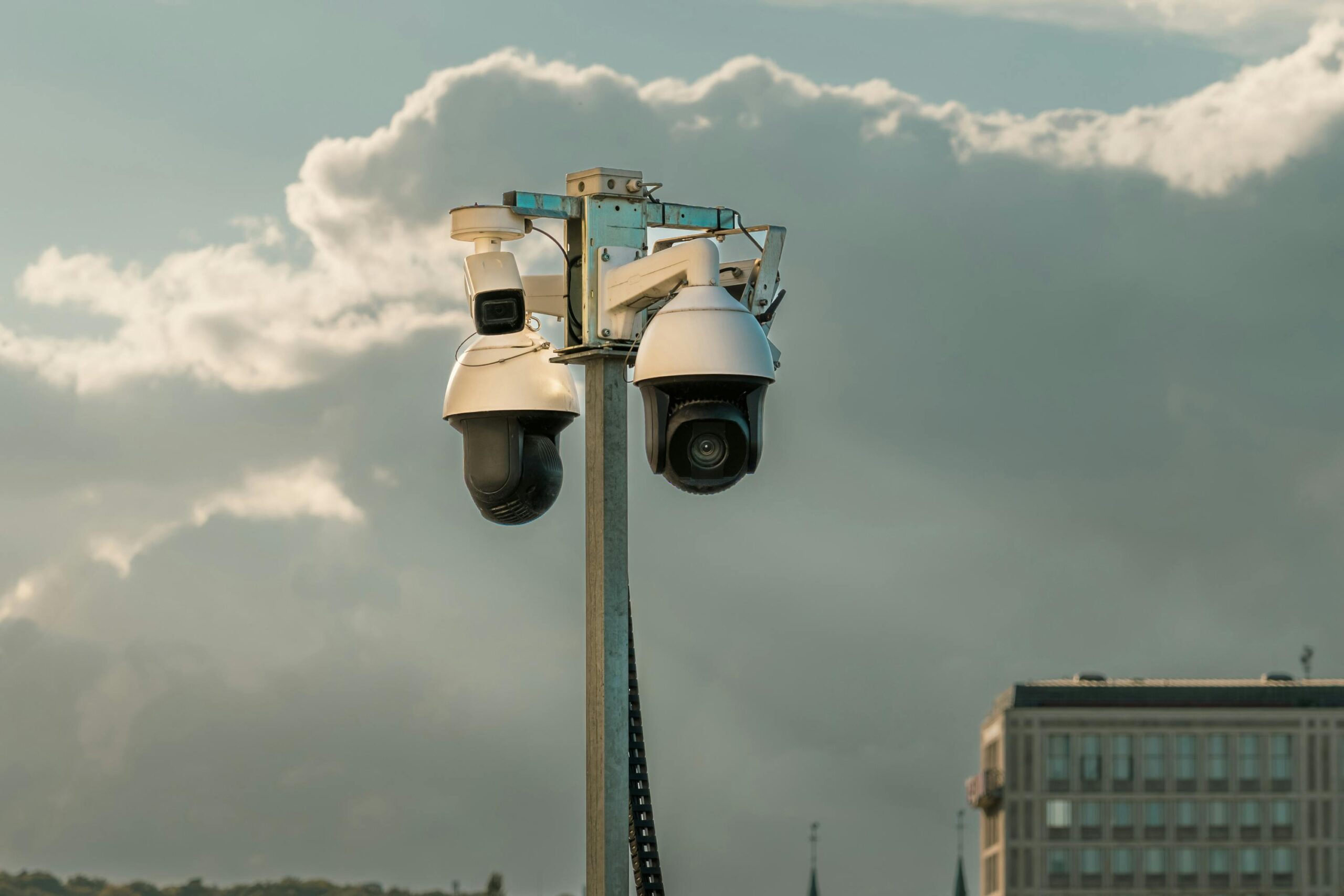 Two modern surveillance cameras mounted on a pole in an urban setting under a cloudy sky.